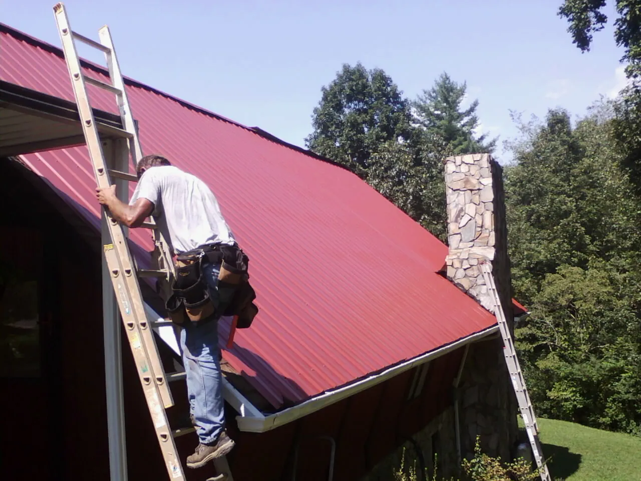 Roofer on a ladder beside a red metal roof and stone chimney — Soffit & Fascia Repair by Roofing Brothers in Howard